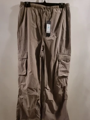 Beige cargopants från 157 Funktion XS - Snygga beige cargopants från 157 Funktion i storlek XS. Byxorna har elastisk midja med snörning och stora fickor på sidorna. Perfekta för en chill och avslappnad stil. Tillverkade i bomullsmaterial för skön känsla.