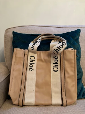 Beige handväska från Chloé - Säljer en snygg beige handväska från Chloé med breda, vita handtag i tyg där loggan står tryckt i svart. Väskan har en boxig form och är tillverkad i skinn med detaljer i textil. Perfekt för dig som vill ha en trendig och rymlig väska med lyxig känsla. Köparen betalar frakten. Köp är köp..