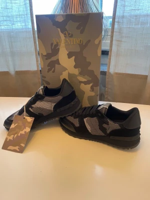 Valentino Garavani Rockrunner sneakers - Säljer ett par Valentino Garavani Rockrunner sneakers i svart, grått och vitt med snyggt kamouflagemönster. Skorna har snörning, meshpaneler och detaljer i skinn och mocka. Kommer med originalkartong, extra skosnören och dustbag.