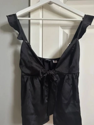 Svart Satinset från Gina Tricot - Säljer ett svart set i glansigt satinmaterial från Gina Tricot. Toppen har breda axelband med volangdetaljer, v-ringning och en söt knytning framtill. Perfekt för dig som gillar en chic och enkel look. Toppen är i storlek M och shortsen storlek S.