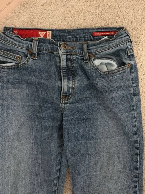 Blå bootcut jeans från Guess - Säljer ett par klassiska blå jeans från Guess med bootcut passform. Har en ikonisk röd etikett i linningen. Defekter finns på bilderna, har spätt upp sömmarna i ändarna vilket ger en snygg detalj. Vintage 