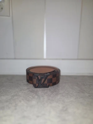 Brun LV-rutig bälte - Säljer ett brunt bälte från Louis Vuitton med klassiskt rutigt mönster och stor LV-logga i metall framtill. Bältet har en stilren design och är tillverkat i ett tåligt material som ger en exklusiv känsla. Perfekt accessoar för att lyfta vilken outfit som helst.