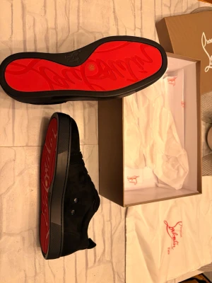 Svarta sneakers från Christian Louboutin - Säljer ett par svarta sneakers från Christian Louboutin i mocka med ikonisk röd sula. Skorna har rund tå, snörning och diskreta metalldetaljer vid sidan. Snygg och stilren design som sticker ut med den klassiska röda undersidan. Dustbag och låda ingår