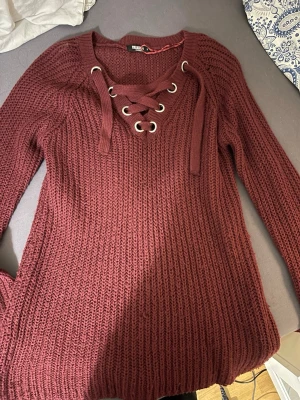 Vinröd stickad tröja med snörning - Mysig vinröd stickad tröja från Colloseum med grov ribbstickning och snygg snörning med metallöljetter vid halsen. Långärmad modell som passar perfekt till höst och vinter. Superskön och trendig att bära till jeans eller kjol.