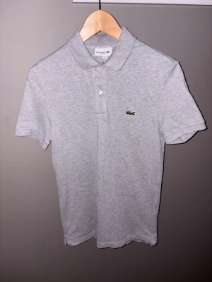 Grå pikétröja från Lacoste - Säljer en klassisk ljusgrå pikétröja från Lacoste med korta ärmar och den ikoniska gröna krokodilloggan broderad på bröstet. Tröjan har krage och knappar framtill. Perfekt till en avslappnad men ändå stilren look.