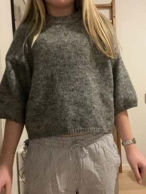 Grå stickad tröja från Gina Tricot - Mysig grå stickad tröja från Gina Tricot i croppad modell. Tröjan har halvlånga ärmar och ribbade detaljer vid halsringning, ärmslut och nederkant. Perfekt för lager på lager under höst och vinter.