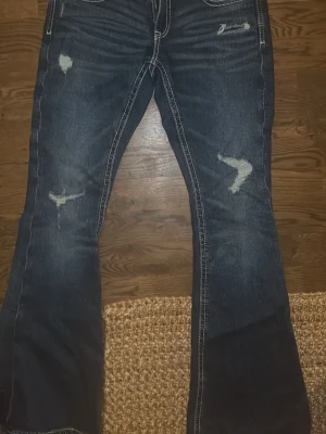 Bootcut jeans från Gina Tricot M - Mörkblå bootcut jeans från Gina Tricot med slitna detaljer och kontrastsömmar. Snygga broderade bakfickor med vit tråd och klassisk femficksmodell. Jeansen har låg midja och är tillverkade i jeansmaterial.