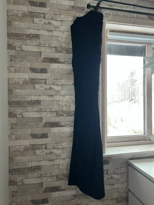 Svart långklänning från Gina Tricot - Säljer en svart långklänning från Gina Tricot i storlek S. Klänningen har en tight passform och är ärmlös med snygg rynkning längs sidan som ger en cool siluett. Perfekt för dig som vill ha en enkel men stilren look.