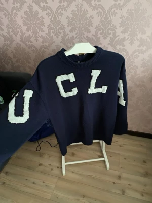 Mörkblå stickad UCLA tröja - Säljer en mörkblå stickad tröja från H&M i samarbete med UCLA. Tröjan har stora vita bokstäver på bröstet och ärmarna, samt en klassisk rund halsringning och lång ärm. Perfekt för dig som gillar college-vibe och oversized känsla.