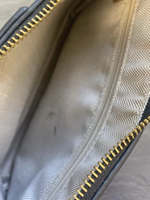 Svart axelväska med kedja - Snygg svart axelväska med klassiskt monogrammönster. Väskan har guldfärgad kedja som axelrem och dragkedja upptill. Insidan är ljus med diskret mönster och har ett innerfack med dragkedja. Perfekt storlek för det viktigaste. Bredd 23 cm , botten 5 cm och längd 15 cm.