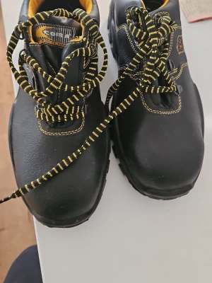 Cofra New Tamigi svarta arbetsskor. Helt nya (aldrig använda) - Säljer ett par svarta Cofra New Tamigi arbetsskor med gula detaljer och snörning. Skorna har robust sula, gul vadderad insida och svart-gula skosnören. Tillverkade i slitstarkt material och designade för superkomfort. Perfekta för dig som vill ha både stil och funktion.