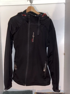 Svart vindjacka från Sail Racing - Svart vindjacka från Sail Racing med huva och snygga röda detaljer vid snörningen. Jackan har dragkedja framtill och två fickor med dragkedja. Materialet är slitstark polyester och jackan har en sportig look med logga på bröstet.