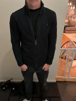 Svart kofta från Polo Ralph Lauren - Säljer en svart kofta från Polo Ralph Lauren i storlek xs. Koftan har ribbad hög krage, hel dragkedja framtill och ett litet rött broderat logomärke på bröstet. Materialet är stickat och ger en skön och stilren känsla. Perfekt för dig som gillar klassisk design. Modellen är 168 cm och väger 55 kg. Det är bara att höra av sig vid frågor och funderingar 😀