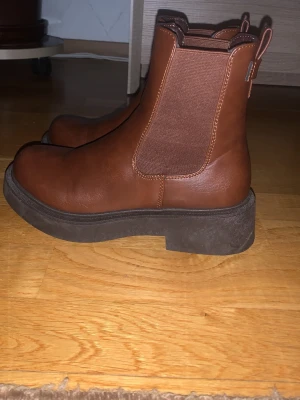 Bruna chelsea boots med platt sula - Snygga bruna chelsea boots i skinn med elastiska paneler på sidorna och grov platt sula i svart. Rund tå och klassisk siluett som passar till många olika stilar. Perfekta för dig som gillar tidlös design och vill ha ett par boots som sticker ut lite extra.