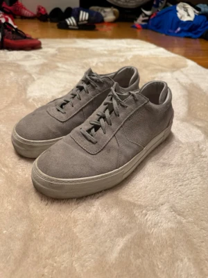 Grå mockasneakers från Axel Arigato - Snygga grå sneakers från Axel Arigato i mocka med klassisk låg profil och vita sulor. Skorna har snörning och en stilren design som passar till det mesta. Perfekta för dig som gillar minimalistisk och clean stil.