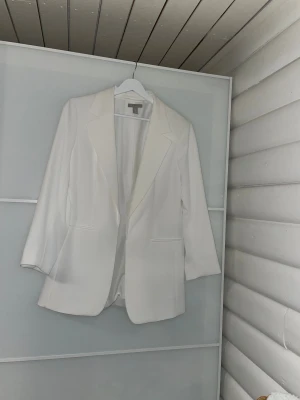 Vit kavaj i storlek 40 - Snygg vit kavaj från H&M i storlek 40. Klassisk modell med långa ärmar, spetsiga slag och två fickor framtill. Tillverkad i mjuk polyester med lite stretch för extra komfort. Perfekt att styla till jeans eller kostymbyxor!