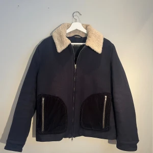 Mörkblå ulljacka med beige krage - Säljer en mörkblå jacka från tiger of sweden med krage i beige. Jackan har två stora fickor i manchester med dragkedjor framtill och quiltat innerfoder. Stängs med dragkedja och har en clean, rak passform. Perfekt för dig som vill ha en stilren och mysig jacka som med tanke på tjocklek funkar lika bra på vintern som vår och höst. Den har en liten reva i innerfodret (se sista bilden). Nypris ca 7000kr