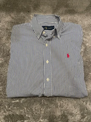 Randig skjorta från Ralph Lauren - Säljer en klassisk skjorta från Ralph Lauren i storlek S. Skjortan är vit med smala marinblå ränder och har en röd broderad logga på bröstet. Tillverkad i bomull och har knappar framtill samt lång ärm. Perfekt för en clean och stilren look.jag är 178 cm och väger 63 kg. Skriv för fler frågor. 