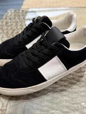 Valentino Garavani  - Snygga sneakers från Valentino Garavani i svart mocka med vita skinnpaneler och vit sula. Skorna har svarta snören och en rund tå. Yttersulan har ett coolt kamouflagemönster och metallplatta med logga. Perfekta för dig som gillar stilrena och lyxiga detaljer. INGE KVITTO LR LÅDA