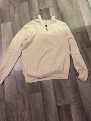 Beige half zip tröja Oscar Jacobson - Beige stickad tröja från Oscar Jacobson med half zip och lång ärm. Klassisk och enkel design, perfekt för lager-på-lager. Tillverkad i mjukt material och har ribbade muddar. Snygg till både jeans och chinos. Ett litet hål vid ena handleden. 