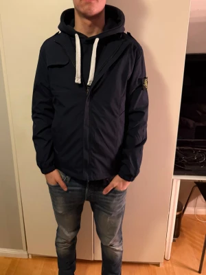 Mörkblå vindjacka från Stone Island - Säljer en mörkblå vindjacka från Stone Island utan huva. Jackan har en klassisk patch på ärmen och är tillverkad i polyester. Snygg och stilren modell som passar perfekt till streetwear. Fodret har coolt medaljmönster.