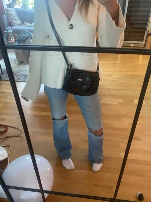 Svart axelväska från Rosetti - Säljer en svart axelväska från Rosetti i mjukt skinnimitation. Väskan har två fickor framtill med lock och vita kontrastsömmar. Praktisk storlek med justerbar axelrem och dragkedja upptill. Perfekt för dig som vill ha en enkel men snygg väska till vardags.