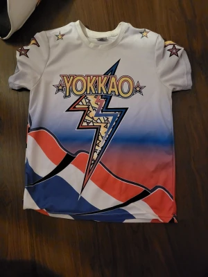 Yokkao t-shirt med blixt och stjärnor - Säljer en fet Yokkao t-shirt i  med korta ärmar. Den har en stor blixt på bröstet, coola stjärnor på axlarna och ett grafiskt mönster i rött, blått och vitt. Perfekt för dig som gillar sportiga och färgstarka prints.