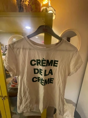 Vit t-shirt med tryck från Holly & Whyte (Lindex) stl L - Säljer en vit t-shirt från Holly & Whyte by Lindex med grön text 'CRÈME DE LA CRÈME' på framsidan. Klassisk rund halsringning och korta ärmar.