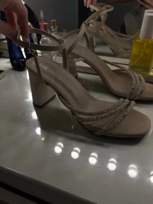Beige sandaler med pärldetaljer från XIT - Säljer ett par beige sandaler från XIT med bred klack och fyrkantig tå. Skorna har tunna remmar dekorerade med små pärlor och glittriga stenar, samt justerbart spänne runt ankeln. Perfekta för dig som vill ha något extra till outfiten.