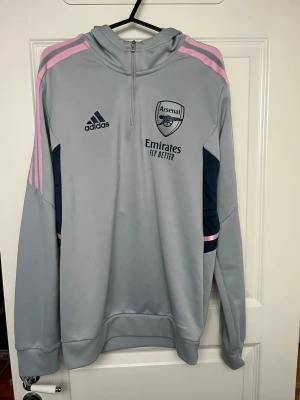 Arsenal hoodie  - Säljer en stilren och bekväm Arsenal hoodie från Adidas i grått med rosa detaljer. Officiell tränings-/uppvärmningströja som användes av Arsenal under säsongen 2021/2022.Tröjan är i  mycket bra skick,utan skador. Perfekt för träning, vardagsbruk eller för dig som är Arsenal-supporter.