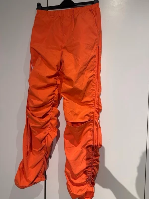 Orange cargopants med snörning - Säljer ett par orange cargopants med coola snörningsdetaljer längs benen som ger en rynkad effekt. Byxorna har elastisk midja och är tillverkade i ett lätt syntetmaterial. Perfekt för dig som vill sticka ut med färg och unik design.