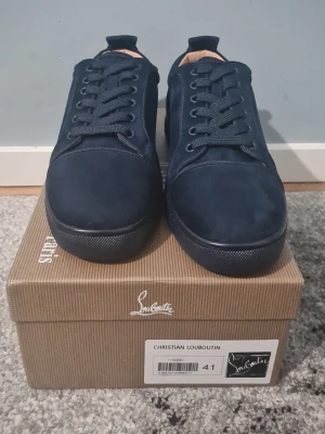 Mörkblå sneakers från Christian Louboutin - Säljer ett par mörkblå sneakers från Christian Louboutin i mocka med klassisk röd sula och broderad logga på hälen. Skorna har rund tå, snörning och en stilren design som sticker ut med den ikoniska röda undersidan.