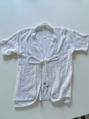 Vit stickad kofta med knytband - Säljer en söt vit stickad kofta med korta ärmar och knytband i midjan. Koftan har ett fint hålmönster och är perfekt att slänga över en topp under varmare dagar. Superfin detalj med små träpärlor på knytbanden. Passar allt från S-L.