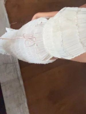 Vit omlottväst med snörning - Säljer en vit topp med snörning i nacken och ribbad struktur. Toppen har volangkant nedtill och är croppad med öppen rygg. Materialet är lätt och känns som syntet, perfekt för varmare dagar. Snygg och trendig design som sticker ut. Säljer även kjolen- köps som set. OBS: kjolen har lite missfärgning nertill, tror att det kan gå bort i en tvätt men vågar inte lova därav pris