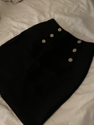 Svart kortkjol med guld knappar - Säljer en svart kortkjol med sex guldiga knappar framtill och en diskret slits baktill. Kjolen har en rak passform och är gjord i ett tjockare tyg som håller formen snyggt. Dragkedja bak för enkel påklädning. 