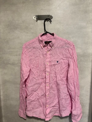 Rosa linneskjorta från Morris - Säljer en rosa skjorta från Morris i 100% linne. Skjortan har klassisk krage, knappar framtill och en liten broderad detalj på bröstet. Perfekt för dig som gillar en avslappnad men ändå stilren look.