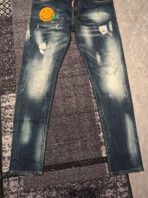 Dsquared2 blå jeans med smiley patch - Säljer ett par blå jeans från Dsquared2 med coolt slitna detaljer och en stor orange smiley-patch på vänster lår. Jeansen har skinny passform, klassisk femficksdesign och är gjorda i jeansmaterial. Färgen är mörkblå med ljusa tvättade partier och slitningar.