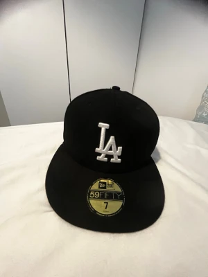Svart LA 59FIFTY keps från New Era - Säljer en svart New Era 59FIFTY keps med LA-logga i vitt framtill. Klassisk flat brim och broderad logotyp. Insidan har märkning och MLB Genuine Merchandise patch. Perfekt för dig som gillar streetwear och sportig stil. Och en klister märke har gåt av 