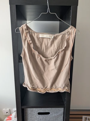 Beige linnetopp från Munthe plus Simonsen - Säljer en beige linnetopp från Munthe plus Simonsen. Toppen har ärmlös design, rundad hals och dekorativa vågiga sömmar längst ner samt ett band som löper längs halsringningen. Perfekt för dig som gillar stilrena och unika detaljer.