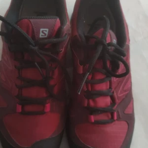 Röda Salomon sneakers med Gore-Tex - Säljer ett par röda Salomon sneakers med svarta detaljer och snörning. Skorna har slitstark yttersula med grovt mönster för bra grepp och är tillverkade i syntetmaterial med Gore-Tex för vattentäthet. Perfekta för utomhusaktiviteter och äventyr.