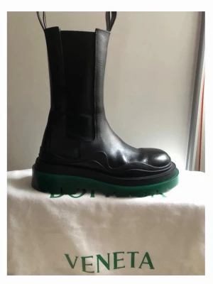Bottega Veneta svarta boots med grön sula - Säljer ett par ikoniska svarta boots från Bottega Veneta med chunky grön sula. Skorna är i skinn med elastiska paneler på sidorna och dragflikar upptill. Riktigt statement-piece med hög plattform och rund tå, perfekta för att lyfta vilken outfit som helst. De är STORA i strl. Så denna 38 är som en 39:a. Nypris 11000kr