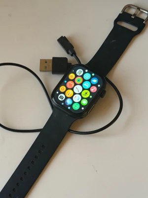 Smart Watch med svart armband - Smart watch med färgskärm och svart silikonarmband. Levereras med laddkabel. Flera appar synliga på skärmen, inklusive hälsa, träning och musik. Nyskick. Perfekt för dig som vill ha koll på aktivitet och notiser direkt på handleden. Passar till Samsung o iPhone. Röda knappar på sidnorna. 