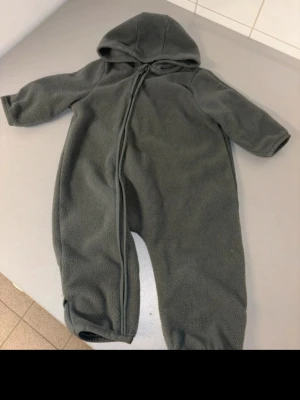 Mörkgrå fleeceoverall med huva - Mysig mörkgrå fleeceoverall med huva och dragkedja som går snett över framsidan. Overallen har långa ärmar och ben, samt vikbara muddar vid händer och fötter för extra värme. Perfekt för kyliga dagar.