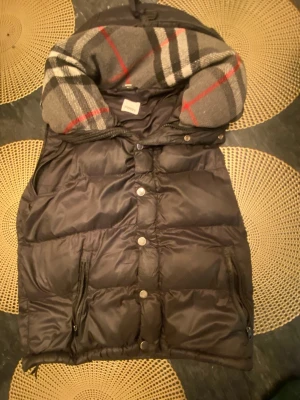 Svart dunväst från Burberry - Säljer en svart dunväst från Burberry med klassiskt rutigt foder i grått, vitt, svart och rött. Västen har tryckknappar, dragkedjor på fickorna och en stor Burberry London England-patch på framsidan. Tillverkad i polyester och dun, perfekt för kyliga dagar.