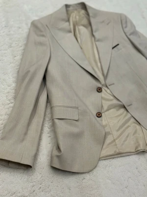 Beige kavaj från Jakmar Classic - Snygg beige kavaj från Jakmar Classic med två bruna knappar framtill och klassiska fickor. Kavajen har ett stilrent snitt, helfodrad insida och diskret mönstrad innerficka. Perfekt för dig som vill ha en clean och tidlös look.