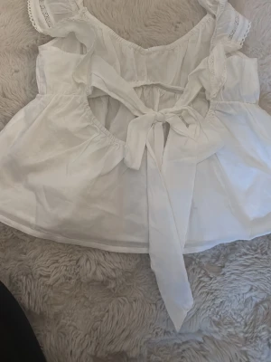 Vit blus med volang och knyt - Supersöt vit blus med volangdetaljer och bred knytning i ryggen. Toppen har ärmlös design med spetskantade axelband och luftig passform. Perfekt för varma dagar och ger en romantisk vibe.