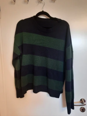 Randig stickad tröja i grönt och blått - Säljer en stickad tröja med breda gröna och mörkblå ränder. Tröjan har rund halsringning och långa ärmar. Perfekt för dig som gillar en chill och avslappnad stil. Passformen är lite loose och materialet känns mjukt och skönt.
