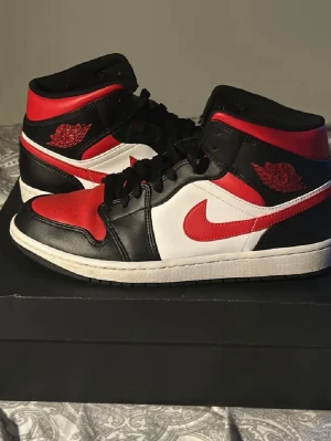 Nike Air Jordan 1 mid röd/svart/vit - Säljer ett par Nike Air Jordan 1 mid med klassisk färgblockning i rött, svart och vitt. Skorna har snörning, rund tå och platt sula. Ikonisk Air Jordan-logga på sidan och röd swoosh. Tillverkade i skinn och kommer med originalkartong. Har använt 1 gång men säljer för att jag sparar pengar