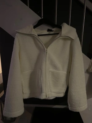 Vit teddyjacka med dragkedja - Mysig vit teddyjacka med stor krage och två stora fickor framtill. Jackan har hel dragkedja och är tillverkad i ett fluffigt teddy-material som ger en chill och trendig vibe. Perfekt för lager-på-lager och att hålla sig varm under kyliga dagar.