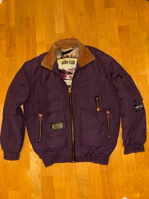 Aero-Club vinterjacka – lila, stl S (44/46) - Snygg och ovanlig jacka från Aero-Club med tydlig flyg-/pilotinspirerad design. Jackan är i en djup lila nyans med kontrasterande dragkedjor i metall och en brun krage. Fodrad insida med dekorativt mönster och stort tryck med flygplansmotiv, vilket ger jackan en unik karaktär.  Flera praktiska fickor med dragkedja framtill samt på ärmen. Resår i midja och ärmslut för bra passform. Patchdetaljer och metallbricka framtill ger ett genomarbetat och robust intryck.  Skick: Mycket bra skick Storlek: S